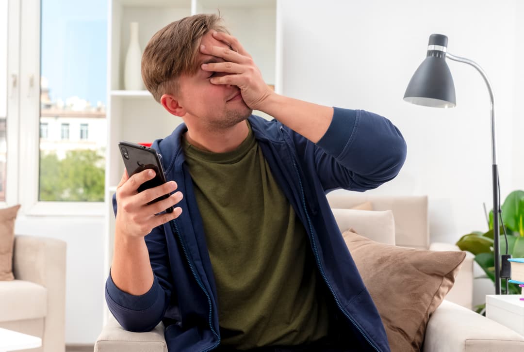 Disappointed young blonde handsome man sits armchair putting hand face holding phone